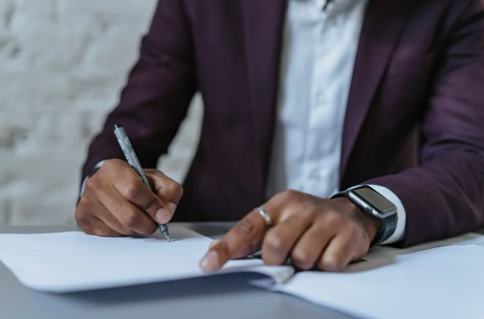 A man writing on white paper with a black pen at a desk.