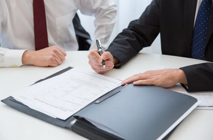 A person in a black suit holding a pen over documents on a desk.