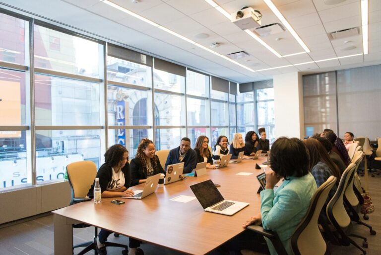 A business meeting is going on with people sitting on a long table with laptops