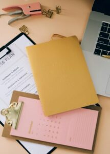 This photo shows a puncher, a clipboard with notes, a laptop, and documents on a desk.