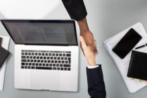 Two people shaking hands with a laptop in the background